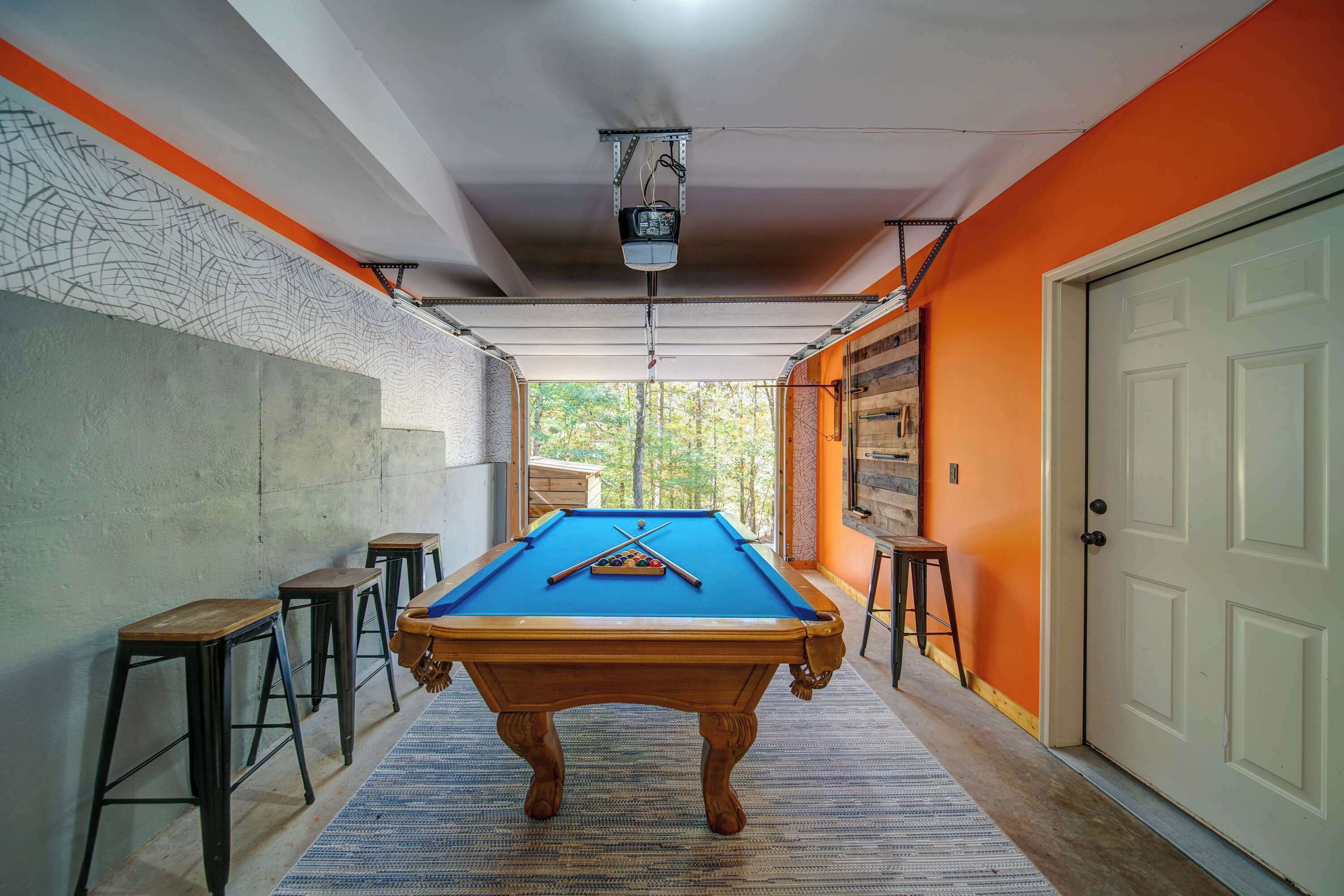 Une table de billard dans un couloir, avec un mur en béton, des tabourets en bois et une porte.