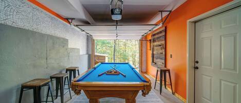 Une table de billard dans un couloir, avec un mur en béton, des tabourets en bois et une porte.