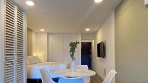 A modern, well-lit room with a bed, a TV mounted on the wall, and a dining area with a table and chairs.