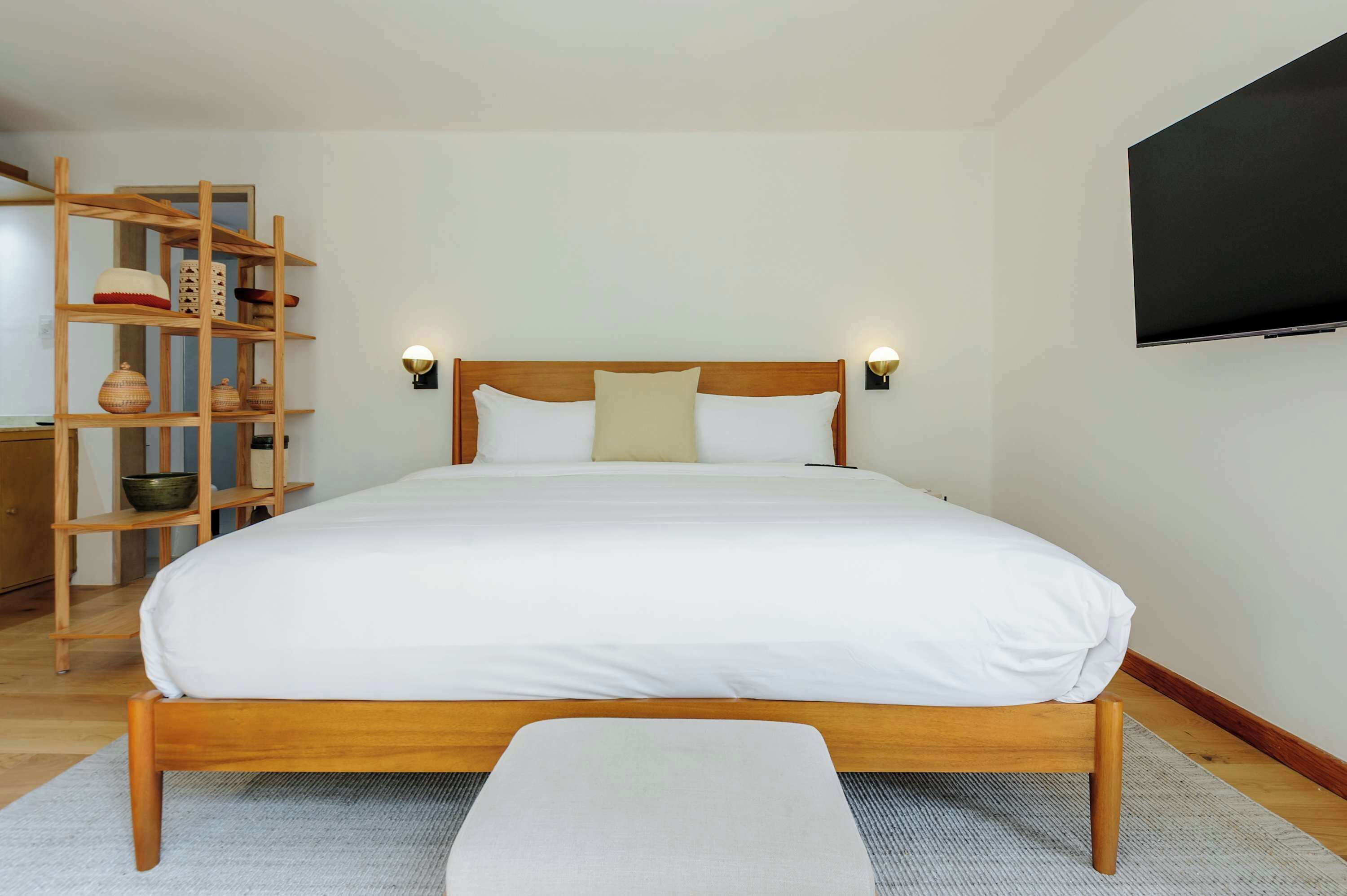A neatly made bed with a wooden headboard, a bedside table, a wall-mounted TV, and a wooden shelving unit with decorative items.