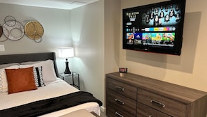 A hotel room with a bed, bedside table, lamp, dresser, and a TV displaying a movie selection. - Cozy Apartment in Superb Fort Washington MD-Private Keyless Entry. (Fort Washington)