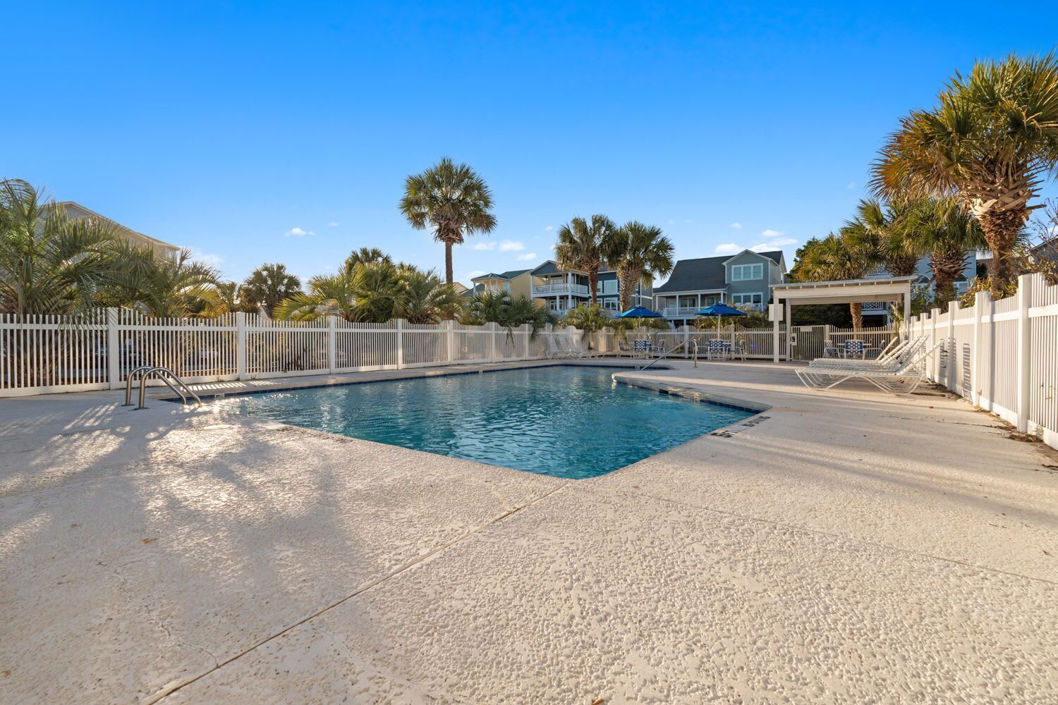 A swimming pool with a white fence, palm trees, and a covered area with lounge chairs.