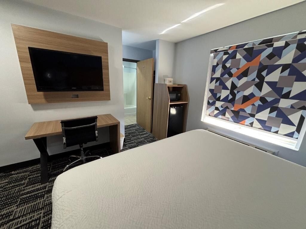 A hotel room with a bed, a desk, a television mounted on the wall, and a window with a geometric pattern.