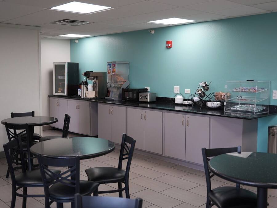 A small, clean kitchenette with a microwave, coffee maker, and a refrigerator. There are two tables with chairs, and a trash can is visible.