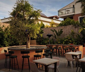An outdoor seating area with tables and chairs, surrounded by plants and a tree.