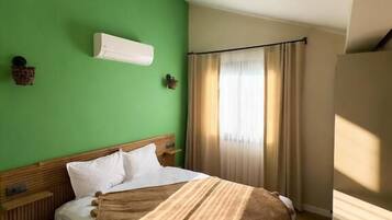 Hotelkamer met een groene muur, een bed met witte lakens, een houten hoofdbord, een nachtkastje en een raam met gordijnen.