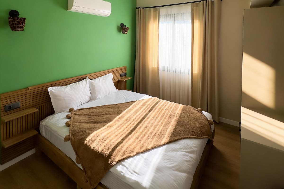 Una habitación con pared verde, cama de madera con ropa de cama blanca, manta marrón y una ventana con cortinas.
