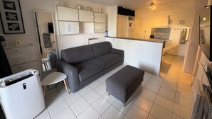 Un espacio habitable compacto con un sofá gris, una mesita pequeña, un armario blanco y una zona de kitchenette.