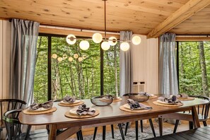 Ein Essbereich mit Holztisch, gedeckt für vier Personen, umgeben von Stühlen und mit Blick auf einen Wald durch große Fenster.