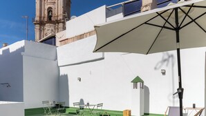 Een terras buiten met een houten tafel en stoelen, een grote parasol en een klokkentoren op de achtergrond.