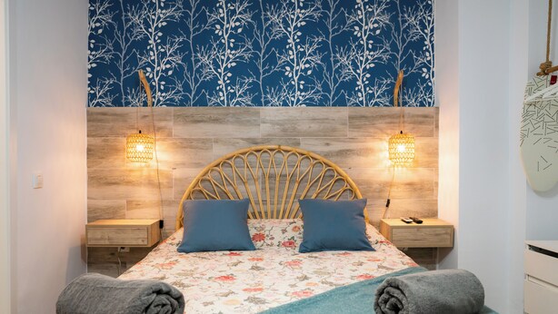 Een bed met een bloemenprint, twee blauwe kussens en een houten hoofdbord. Er zijn twee nachtkastjes met lampen en een wandklok.