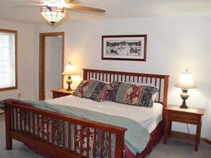 Une chambre à coucher avec un lit en bois, deux tables de chevet avec des lampes, un ventilateur de plafond, un tableau encadré au mur et une porte.
