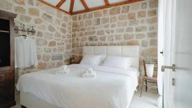 Une chambre avec un mur en pierre, un plafond en bois, un grand lit avec une literie blanche, une table de chevet et une chaise agrémentée d’une plante en pot.