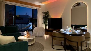 En moderne stue med sofa, rundt sofabord, spiseplads med stole, fjernsyn og en balkon med udsigt.