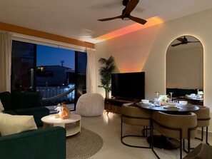 Ein modernes Wohnzimmer mit einer Couch, einem runden Couchtisch, einem Essbereich mit Stühlen, einem Fernseher und einem Balkon mit Blick.