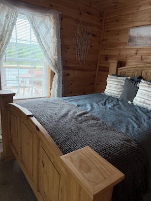 A wooden cabin bedroom with a bed, curtains, a dreamcatcher, and a painting on the wall. - Harmony Cove - Peaceful, serene 2-bdrm cabin on the lake in charming Tunnel Hill (Tunnel Hill)
