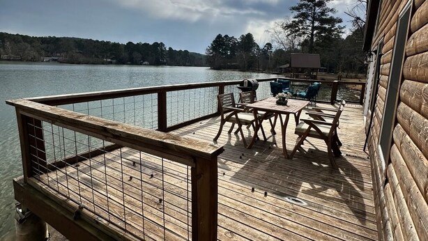 En træterrasse med gelænder og udsigt over en sø, med et bord, stole og en grill.
