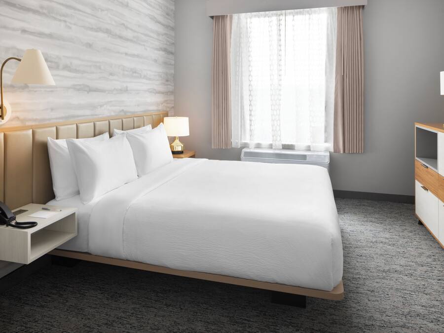 Ein Hotelzimmer mit einem Bett, Nachttischen, einer Lampe, einem Telefon, einem Fenster mit Vorhängen und einer Wand mit strukturierter Oberfläche.