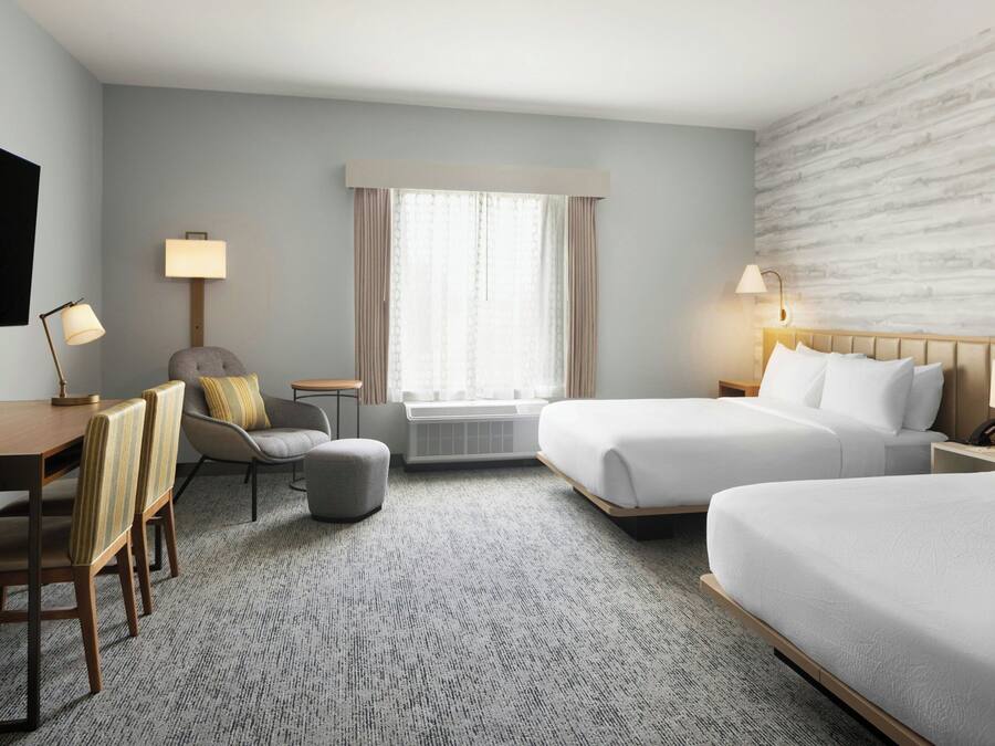 A hotel room with two beds, a desk, a chair, a small table, and a window with curtains.