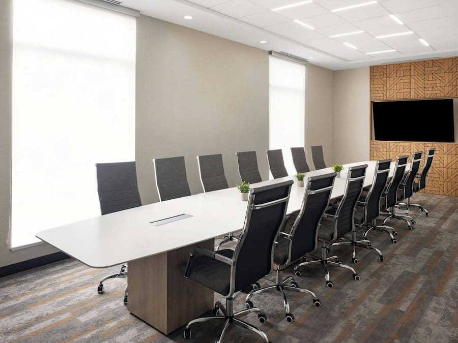 Un salón de conferencias con una larga mesa, sillas negras, un televisor en la pared y ventanas grandes.