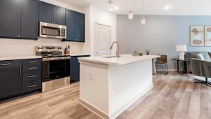 A modern kitchen with dark cabinets, a stainless steel oven, a microwave, and a white island with a sink. - Landing | Stunning 1BD, Gym, Yoga Studio (Huntsville)