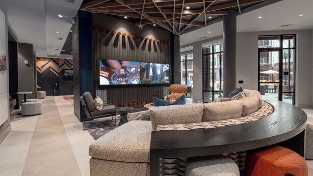 A modern living room with a curved sectional sofa, a flat-screen TV mounted on the wall, and a large window providing a view of the outdoors. - Landing | Stunning 1BD, Gym, Yoga Studio (Huntsville)