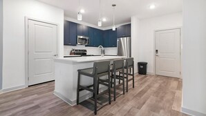 A modern kitchen with dark blue cabinets, white countertops, a stainless steel refrigerator, a microwave, and a sink. There are three bar stools and a trash bin. - Landing | Spectacular 1BD, Yoga Studio, Gym (Huntsville)
