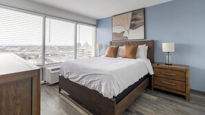 A modern bedroom with a large bed, wooden headboard, two bedside tables with lamps, and a city view through floor-to-ceiling windows. - Landing | Amazing 1BD, Game Room, Yoga Studio (Hartford)