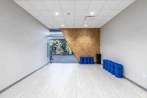 A modern hallway with a wooden wall panel, a glass door leading to a gym area, and blue cylindrical objects on the floor. - Landing | Beautiful 1BD, Game Room, Gym (Hartford)