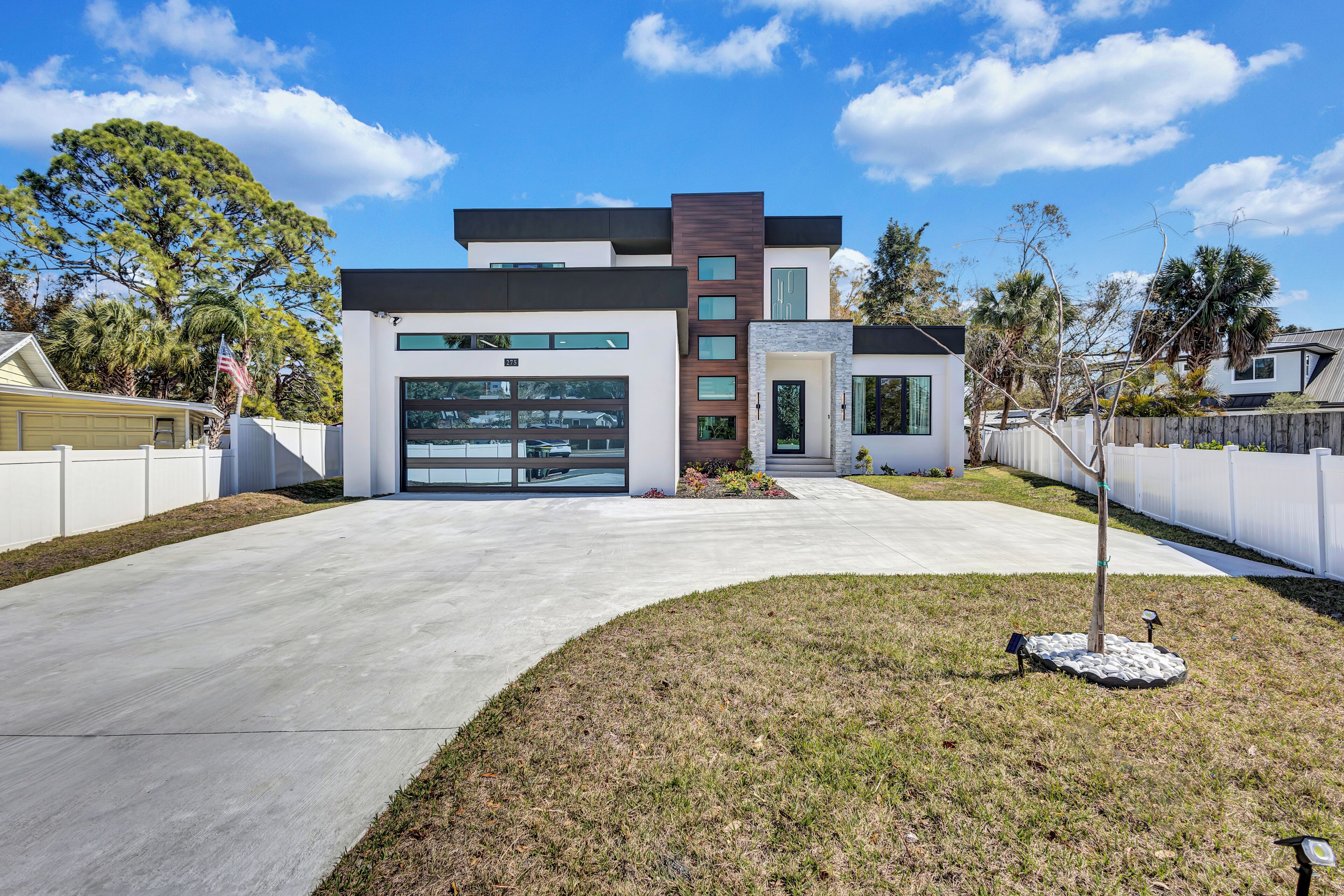 A modern two-story house with a garage, a front yard with a small tree, and a white fence.