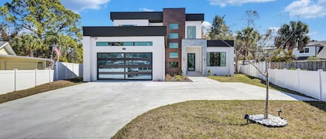 A modern two-story house with a garage, a front yard with a small tree, and a white fence.