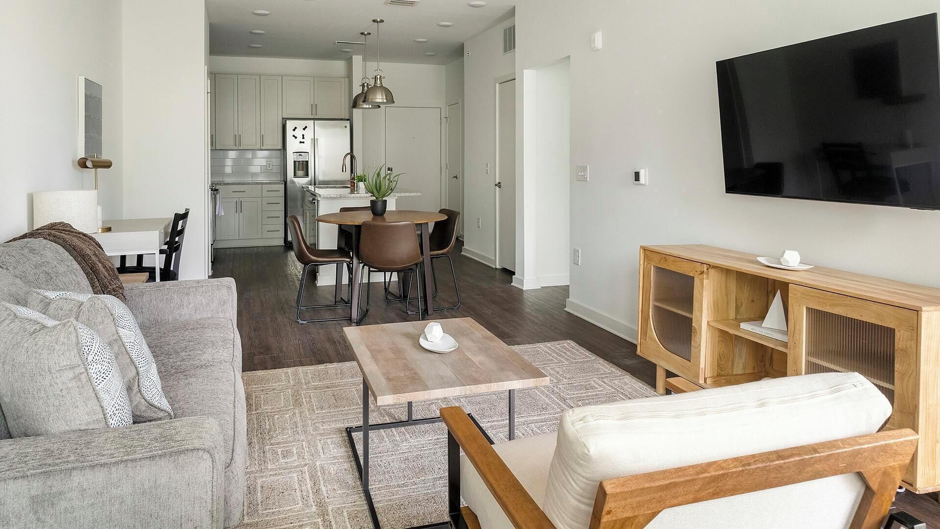 A modern living room with a sofa, coffee table, and TV stand. There is a dining area with chairs and a table. The kitchen is visible in the background.