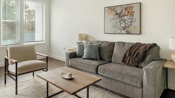 A living room with a grey sofa, a wooden coffee table, a beige armchair, and a painting on the wall. - Landing | Chic 1BD, Clubhouse, Yoga Studio (Raleigh)