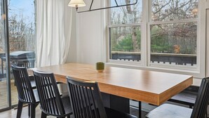 Un comedor con mesa de madera y sillas negras, ventanas amplias con vista a árboles y una terraza.