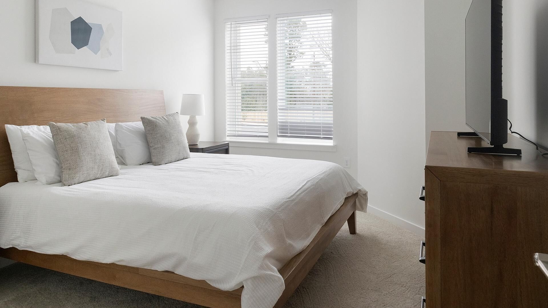 Ein Schlafzimmer mit Bett, Nachttisch, Lampe, Fernseher und einem Fenster mit Jalousien.