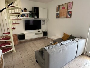 Un salon avec un canapé gris, un téléviseur, un escalier blanc et une bibliothèque.