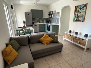 Una sala moderna con un sofá seccional gris, una mesa de comedor con sillas, una cocina con armarios azul claro y un aparador blanco con frascos.
