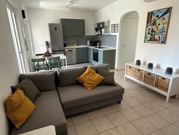 Un salon moderne avec un canapé d’angle gris, une table à manger avec des chaises, une cuisine avec des meubles bleu clair et une console blanche ornée de vases.