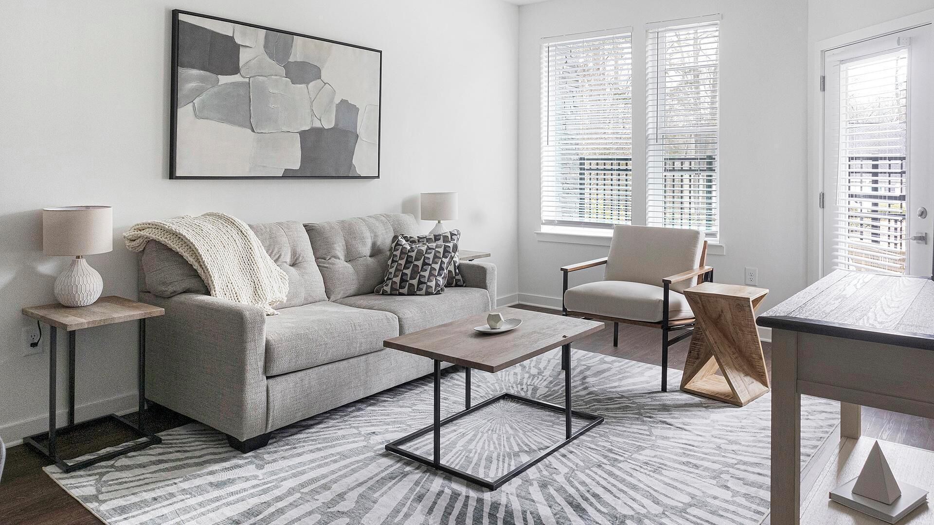 Una sala moderna con un sofá gris, una mesa de centro, una mesita auxiliar con una lámpara, una silla y un cuadro abstracto grande en la pared.