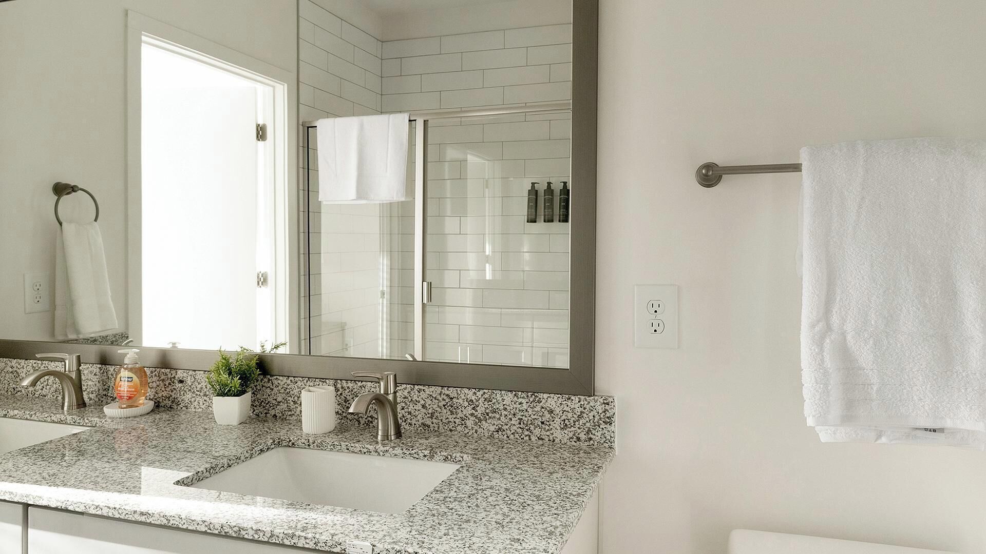 Une salle de bain dotée d’un comptoir en granit, de deux lavabos, d’un grand miroir, d’un porte-serviettes et d’un espace douche aux carreaux blancs.