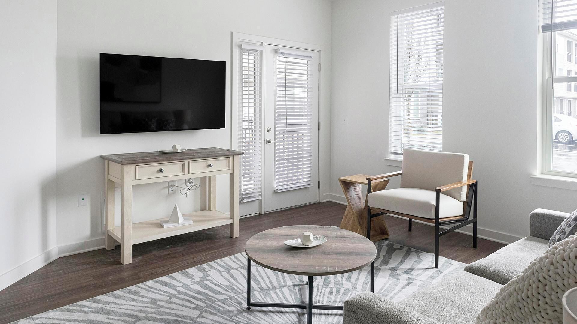 A modern living room with a flat-screen TV, a wooden side table, a round coffee table, a beige armchair, and a grey sofa.