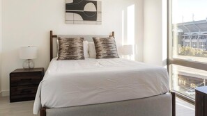 A neatly made bed with a white comforter and two patterned pillows, a bedside table with a lamp, and a large window with a view of a building and trees. - Landing | Amazing 1BD in Uptown Charlotte (Charlotte)