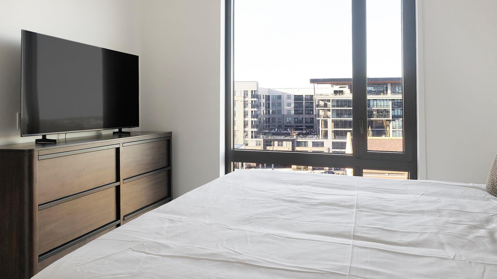 Een slaapkamer met een bed, een dressoir, een televisie en een groot raam met uitzicht op de stad.