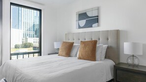 Habitación de hotel con una cama grande, dos lámparas de noche, vistas de la ciudad a través de una ventana y un cuadro abstracto moderno en la pared.