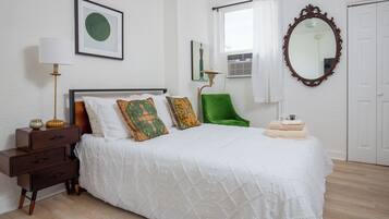A neatly arranged bedroom with a bed, a bedside table, a lamp, a chair, and a mirror.