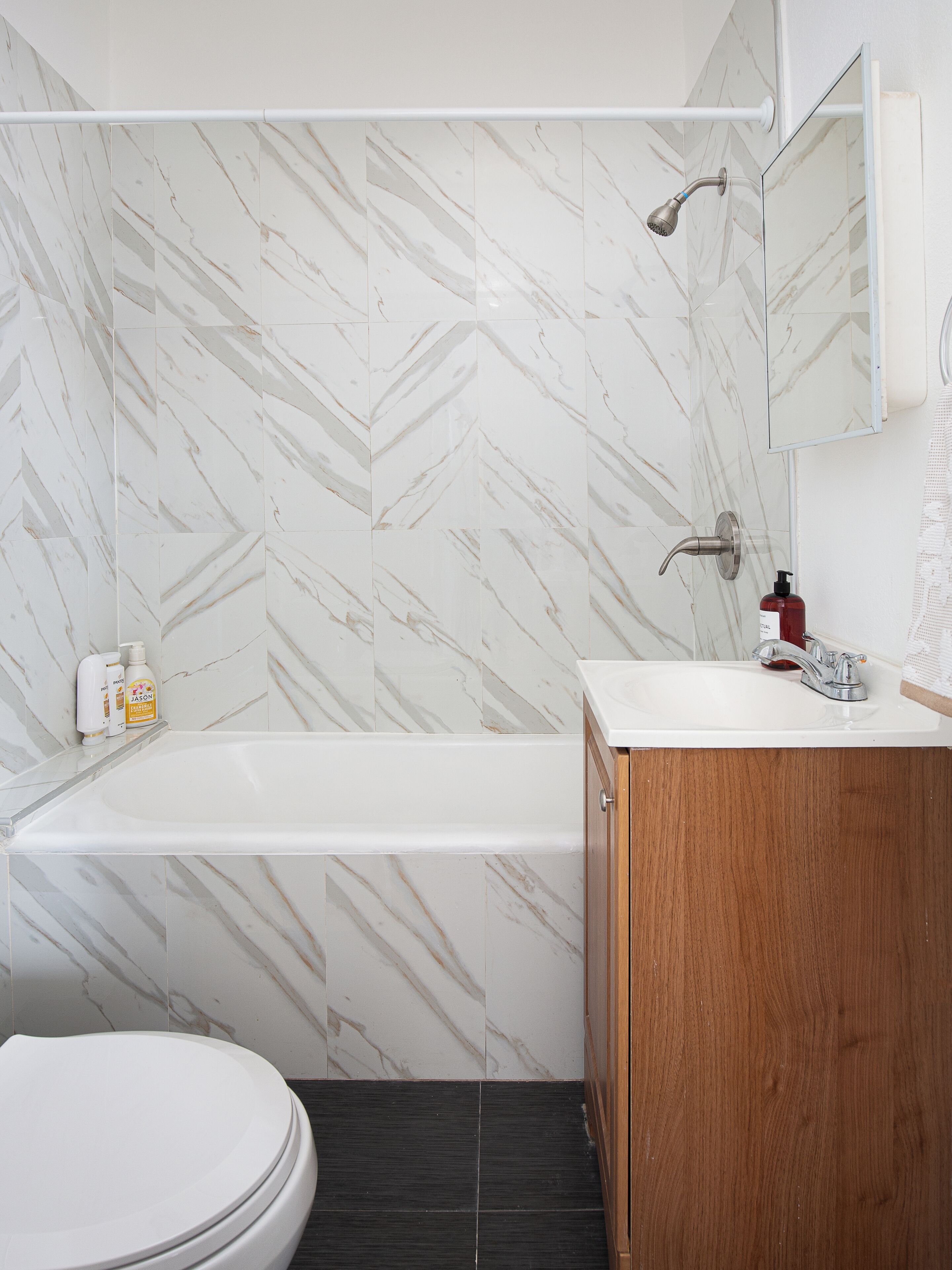 A bathroom with a marble-patterned wall, a white bathtub, a wooden vanity, and a toilet.
