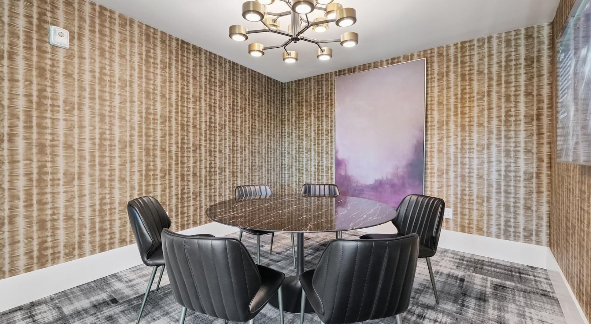 Een vergaderruimte met een ronde tafel, zes zwarte lederen stoelen, een moderne kroonluchter en behang met een patroon.