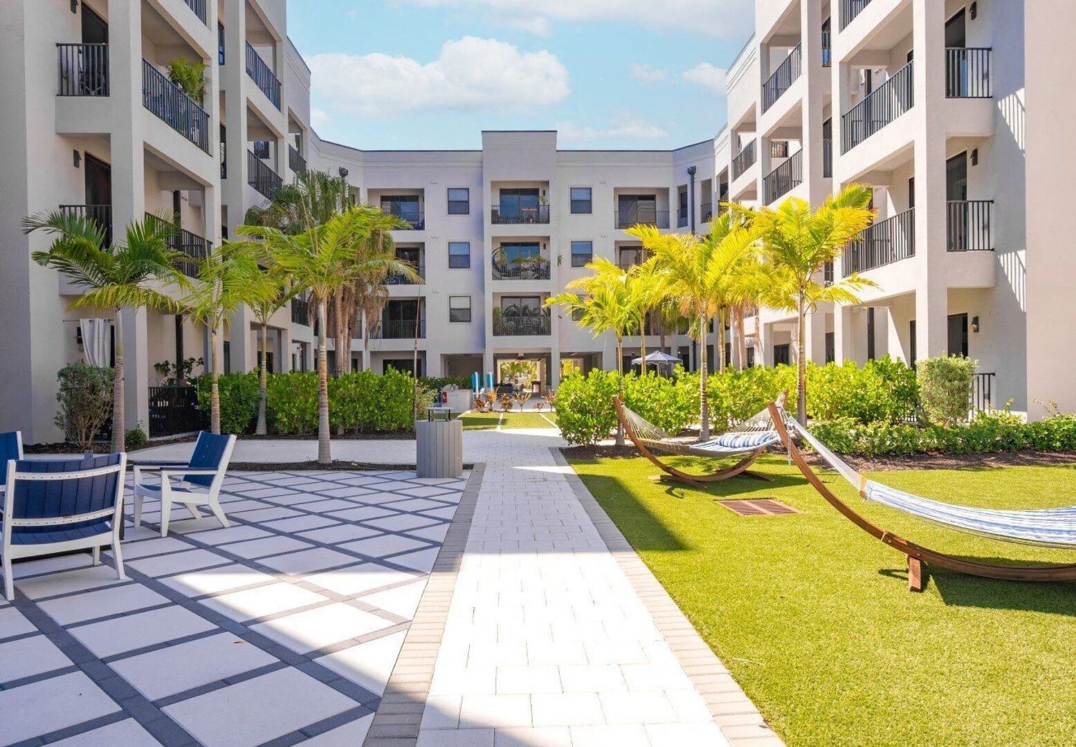 A modern multi-story residential building with balconies, a paved walkway, and a well-maintained garden area with palm trees and a hammock.