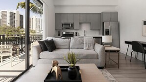 Eine moderne Wohnung mit grauem Sofa, einem Essbereich, einer Küche mit grauen Schränken und Blick auf Palmen und Gebäude.