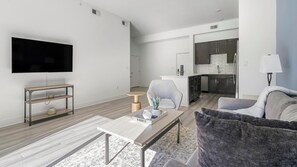 Ein modernes Wohnzimmer mit Flachbildfernseher, Couch, Couchtisch und einer Küchenzeile inklusive Schränke und Geschirrspüler.